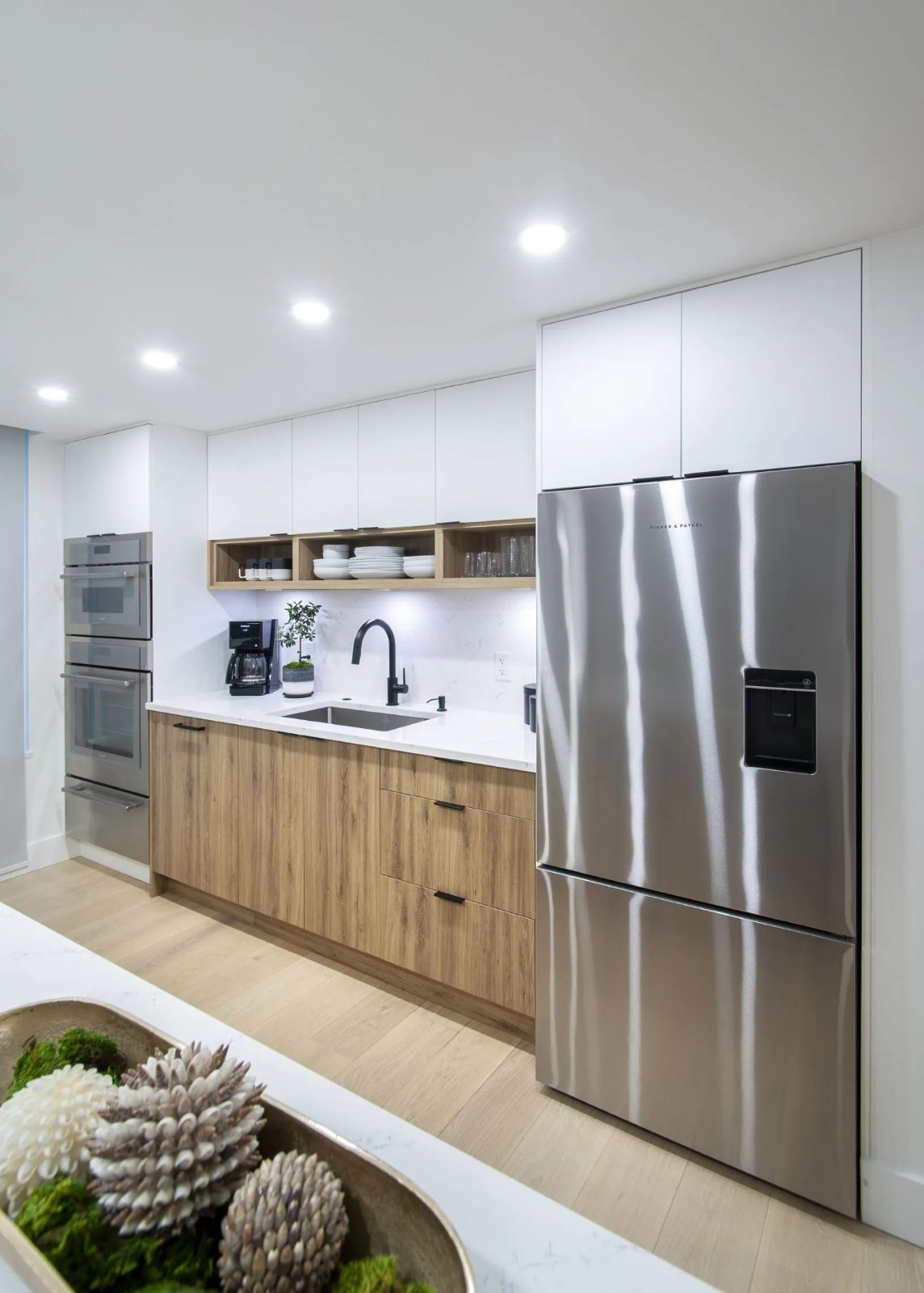 Compact condo kitchen with white uppers to the ceiling, wood-grain lower cabinets, an open oak shelf and a stainless fridge